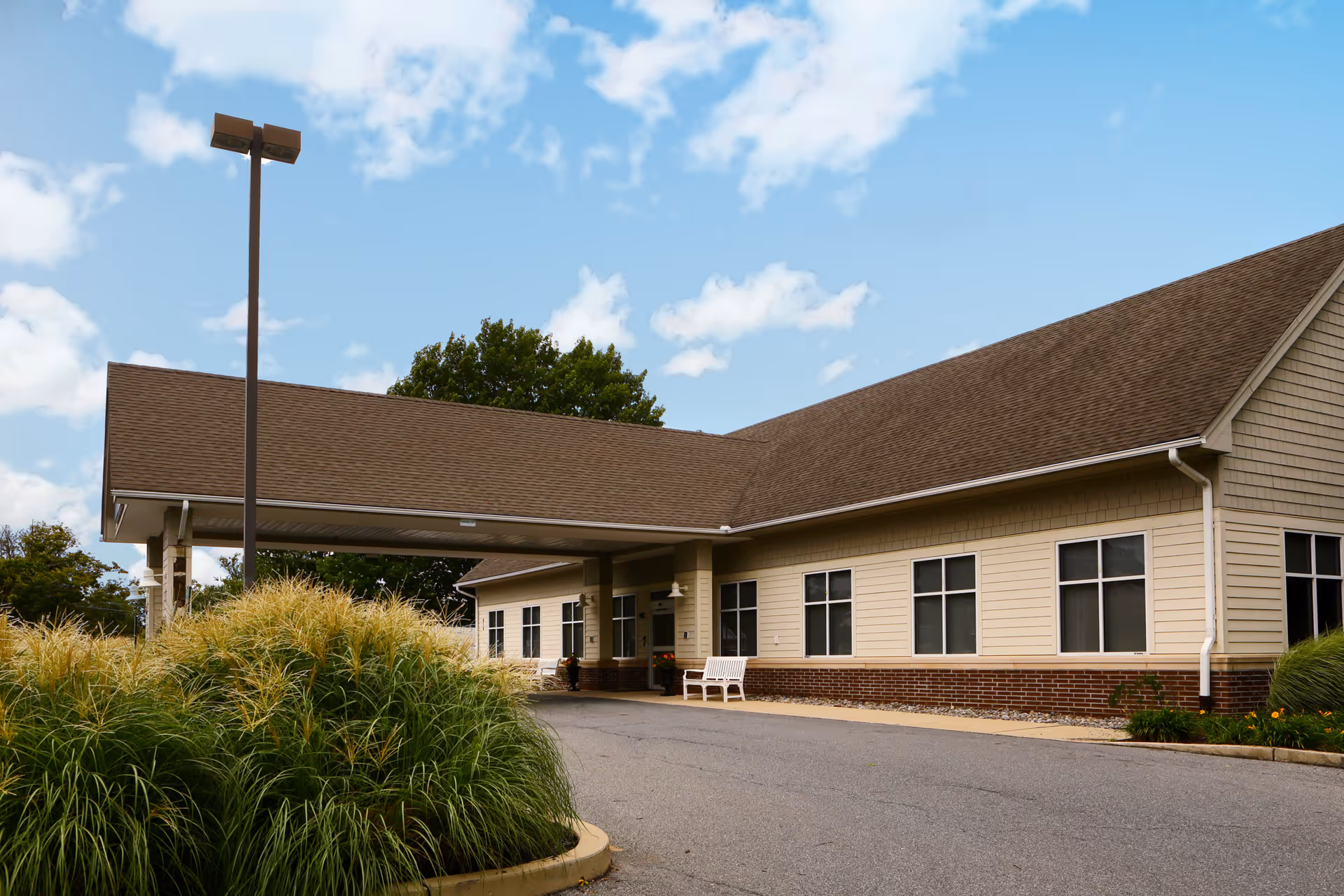 Exterior view of a single-story building with beige siding and a brown roof under a blue sky with scattered clouds. There is a covered entrance with a driveway, a white bench near the entrance, and tall ornamental grasses in the foreground.