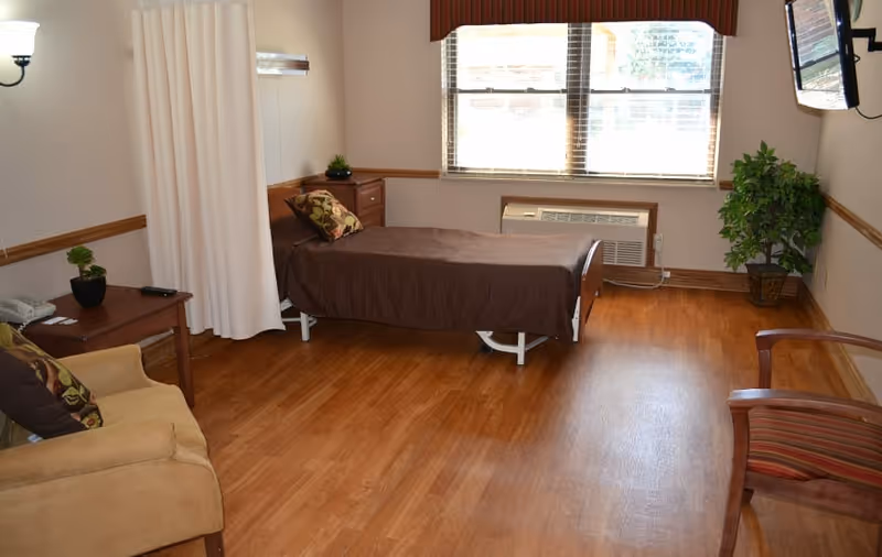 A senior living facility room with a single bed covered in a brown blanket, a wooden nightstand with a small plant, a beige armchair with a patterned pillow, a wooden chair with red and brown striped upholstery, a large window with blinds, a wall-mounted TV, and a potted plant in the corner. The room has wooden flooring and light-colored walls with wooden trim.
