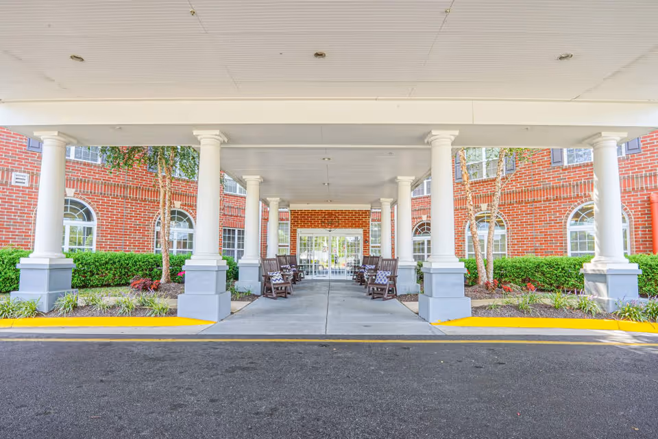 Covered entrance with white columns and rocking chairs leading to glass doors of a red-brick senior living building.