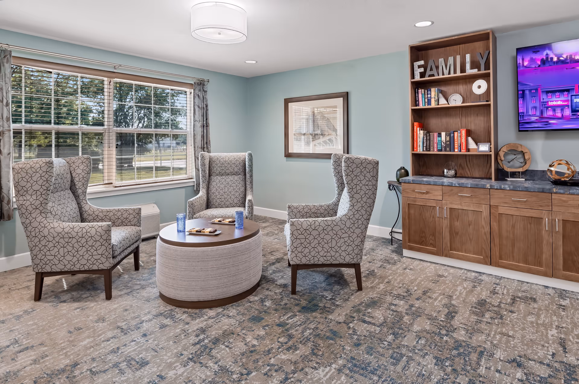 A cozy sitting area in a senior living facility with three patterned armchairs arranged around a round coffee table holding two blue glasses and plates with snacks. The room has a large window with curtains, a framed picture on the wall, and a wooden cabinet with shelves containing books and decorative items, including large letters spelling FAMILY. A flat-screen TV is mounted on the wall above the cabinet.