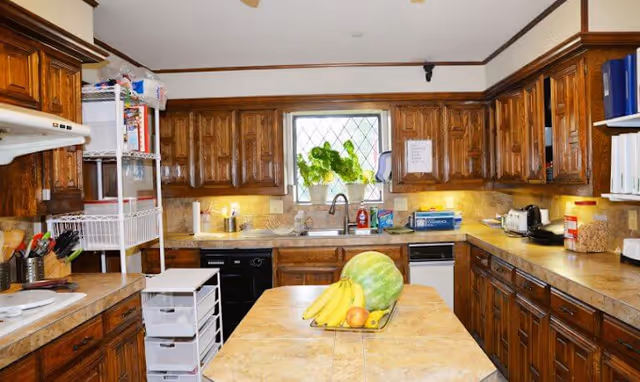 A spacious kitchen with wooden cabinets and beige countertops. There is a central island with a tiled surface holding a tray of fruit including bananas, a watermelon, and an apple. The kitchen features a window above the sink with green plants on the windowsill, a dishwasher, a toaster, and various kitchen utensils and supplies on the counters.