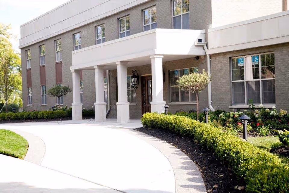 Covered entrance and portico of a brick senior living building with columns, windows, and a landscaped curved driveway.