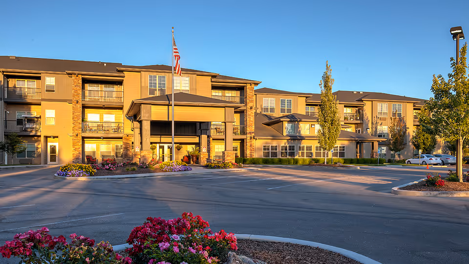 Exterior view of MorningStar Senior Living of Boise building during daylight with a clear blue sky. The three-story building features balconies, large windows, and a covered entrance with an American flagpole in front. There are landscaped flower beds and trees around the parking area.