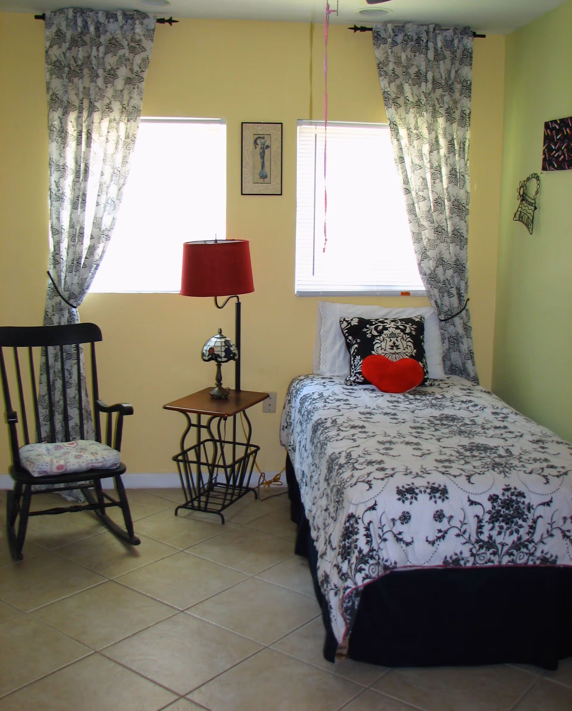 A small bedroom with a single bed covered in a white and black floral patterned bedspread. There is a red heart-shaped pillow and a black and white decorative pillow on the bed. Next to the bed is a small wooden side table with a red lampshade and a small decorative lamp. A black rocking chair with a floral cushion is positioned near two windows with white and black floral curtains. The walls are painted yellow and light green, and there are a few pieces of wall art.