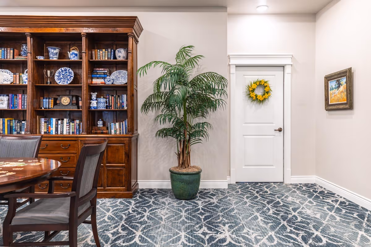 Interior room with a wooden bookshelf filled with books and decorative blue and white ceramics, a round wooden table with chairs, a large potted plant, a white door adorned with a yellow flower wreath, and a framed painting on the wall.