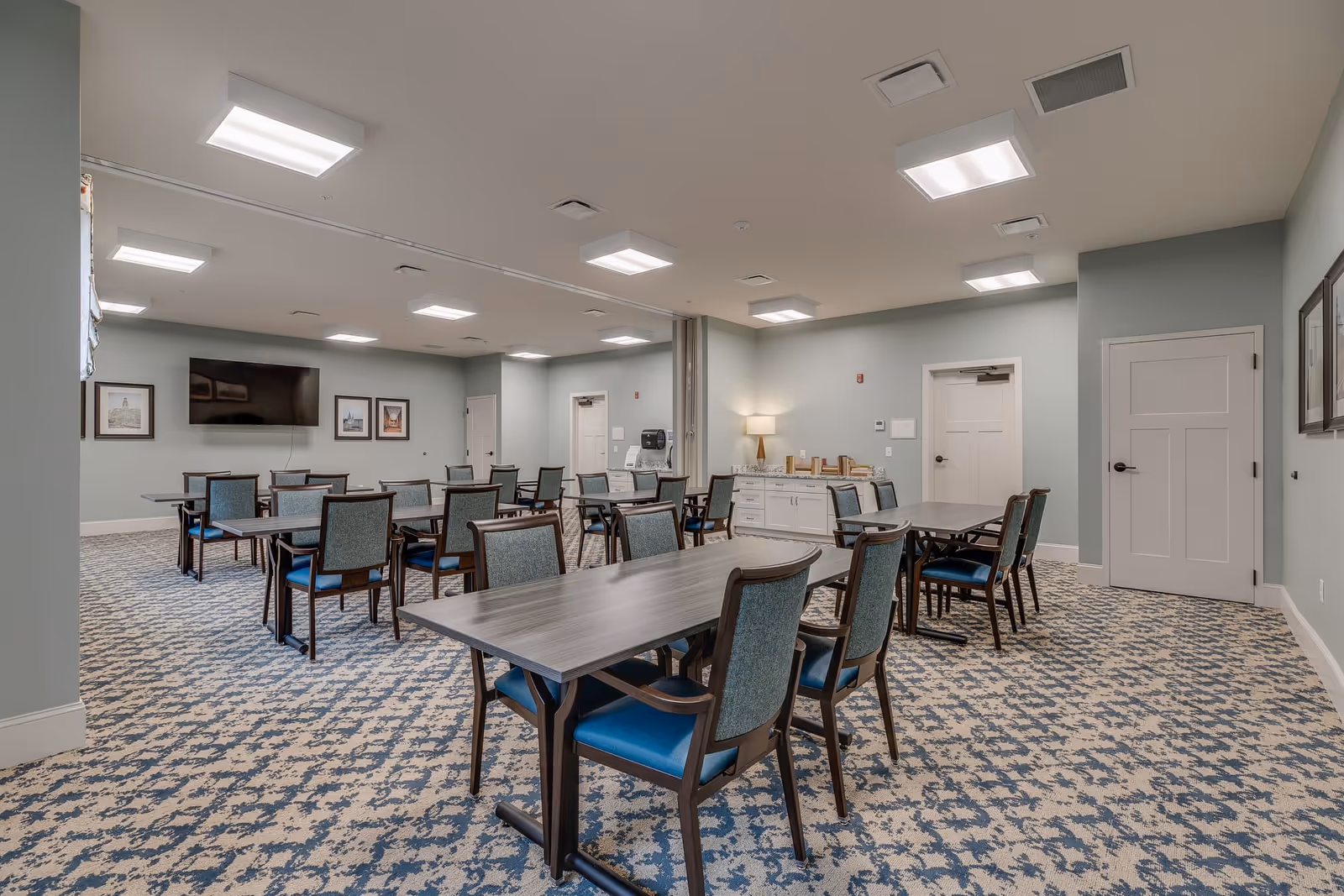 A spacious dining room with multiple rectangular tables and chairs arranged neatly. The room has light blue walls, patterned carpet flooring, and bright ceiling lights. There is a large flat-screen TV mounted on the wall, framed pictures, a sideboard with a lamp, and two closed white doors.