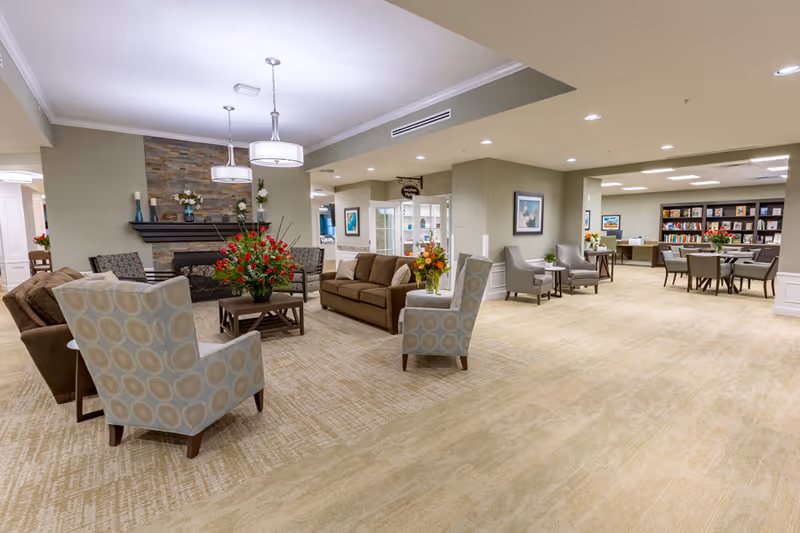 Spacious, well-lit senior living common area with armchairs and sofas arranged around a central coffee table and fireplace, with dining and library seating visible in the background.