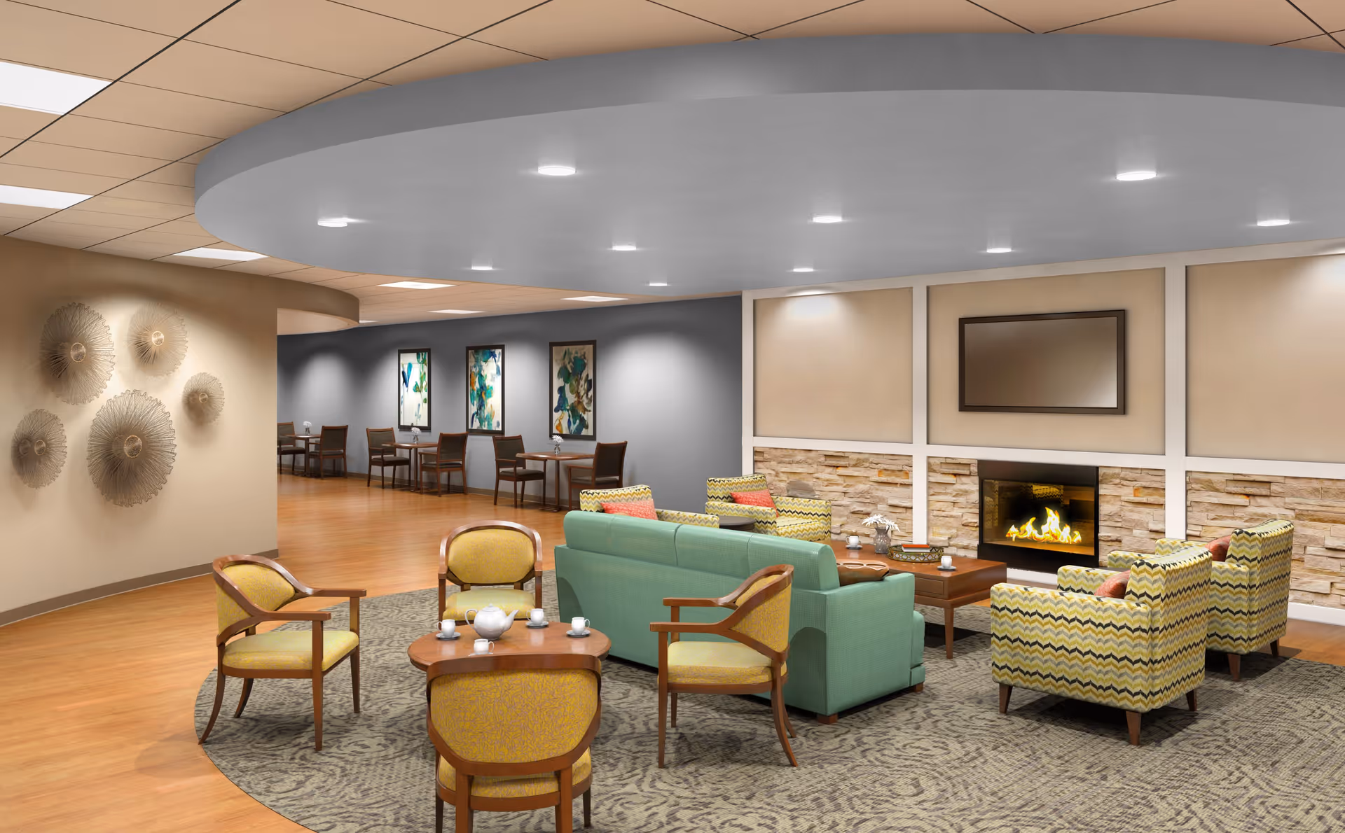 A cozy senior living facility common area with a green sofa, four patterned armchairs, and a wooden coffee table arranged on a large patterned rug. A fireplace with a stone surround and a mounted TV above it is on the far wall. The room has warm lighting, decorative wall art, and a dining area with tables and chairs in the background.