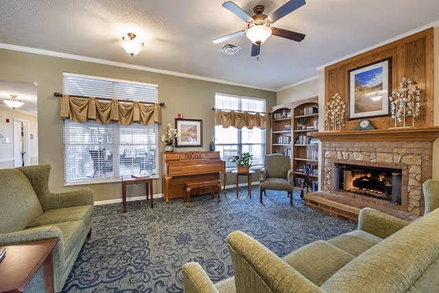 Cozy common room with green sofas and chairs, a piano, built-in bookshelves, and a stone fireplace under a ceiling fan.