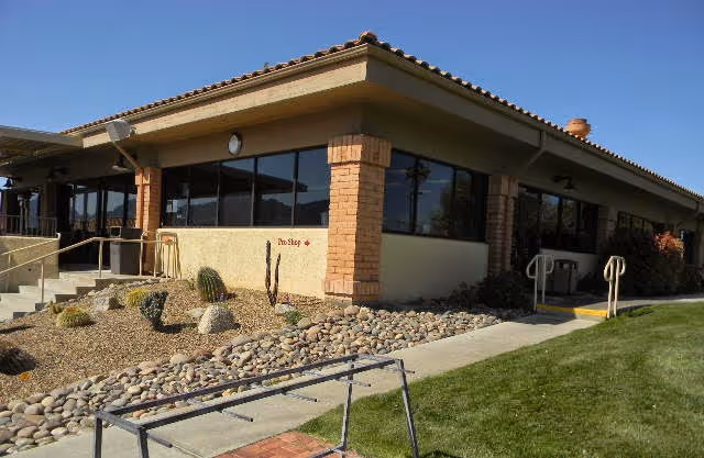 Exterior view of a single-story building with large windows, brick pillars, and a tiled roof. The building is surrounded by a landscaped area with rocks, small desert plants, and a grassy lawn. There are concrete walkways and metal handrails near the entrances.