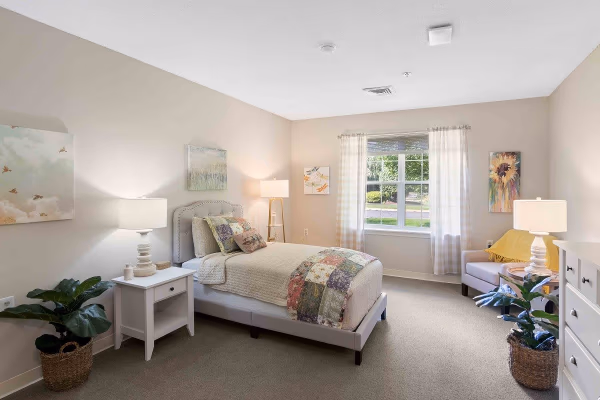 A bright and cozy bedroom with a single bed featuring a patchwork quilt and multiple pillows. The room has beige walls and carpet, a window with sheer curtains letting in natural light, a white nightstand with a lamp and small decor, a white dresser, a comfortable armchair with a yellow throw blanket, two potted plants in woven baskets, and several colorful paintings on the walls.