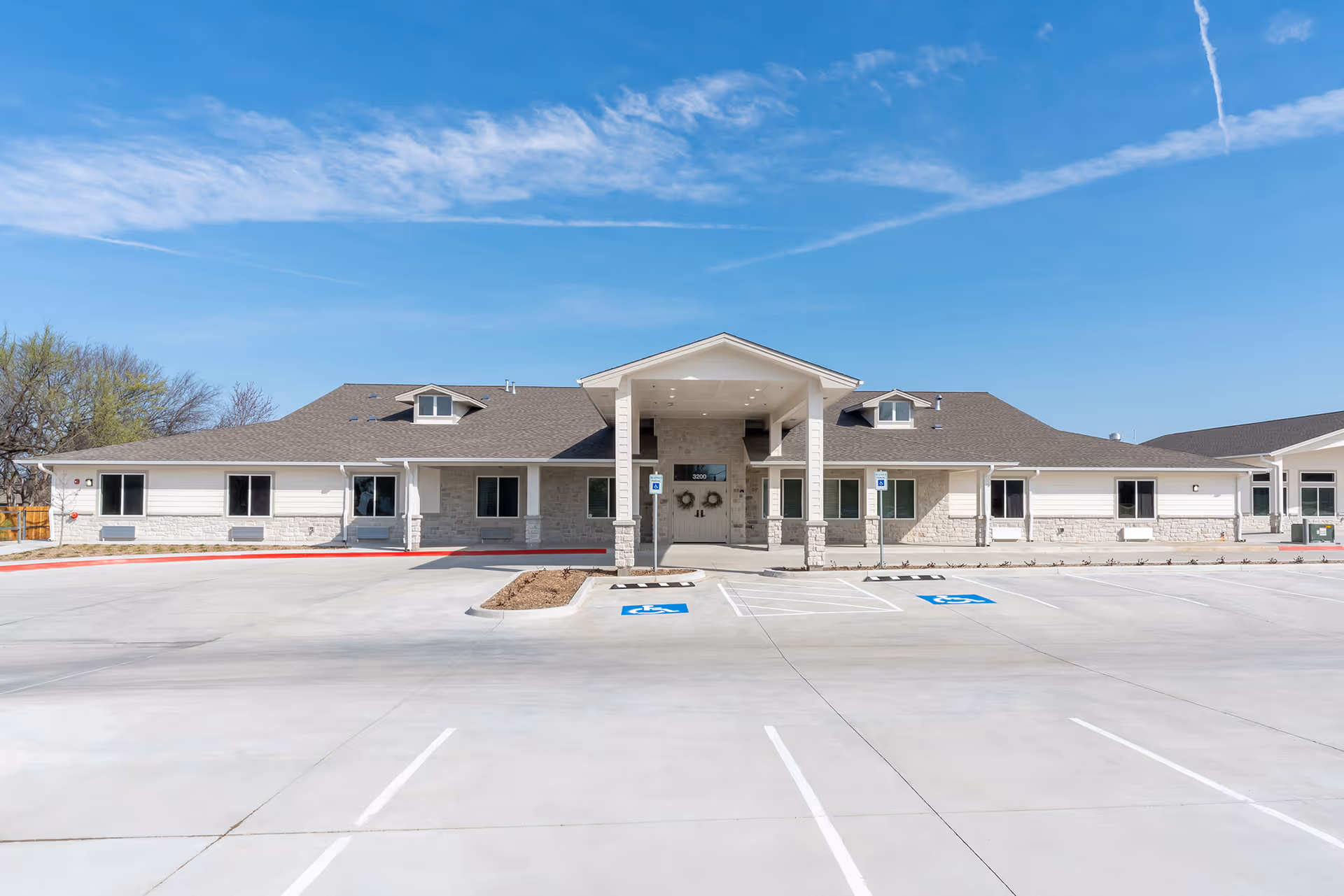 Front exterior view of a single-story senior living facility building with a covered entrance, multiple windows, and a large empty parking lot with marked handicap parking spaces under a clear blue sky.