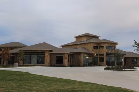 Exterior view of a single-story building with stone and brick facade, large windows, and a multi-level roof under a cloudy sky. The building is surrounded by a paved driveway and some grass with small trees.