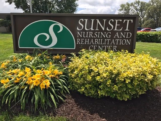 Outdoor sign for Sunset Nursing and Rehabilitation Center surrounded by yellow flowers and green bushes with trees and a cloudy sky in the background.
