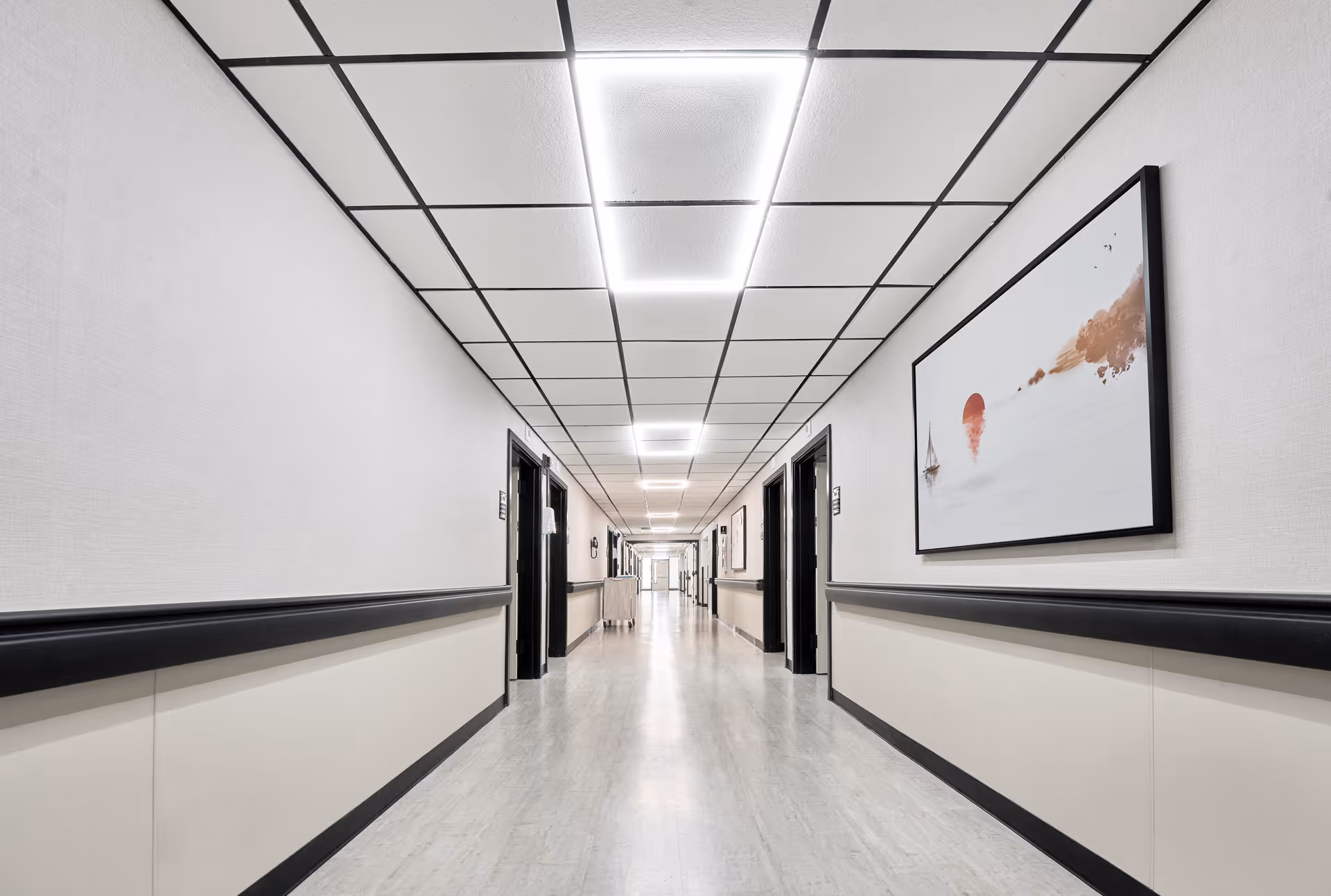 A long, clean hallway in a health and rehabilitation center with white walls, black handrails, and several doorways on both sides. The ceiling has a grid pattern with bright rectangular lights. There are framed artworks on the walls and a cart is visible in the distance.