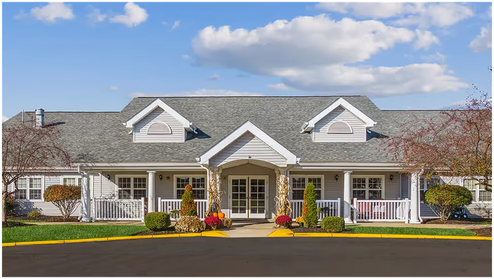 Front exterior view of a single-story senior living facility building with a gray roof, white siding, and a covered entrance with columns. There are small bushes and trees on either side of the entrance, and the sky is partly cloudy.