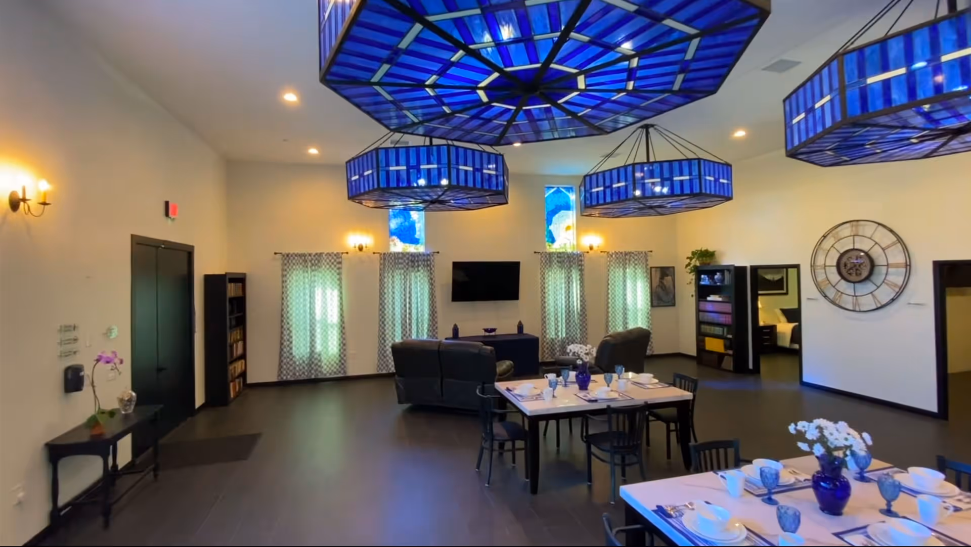 A spacious common area in a senior living facility featuring large blue stained glass pendant lights hanging from the ceiling. The room includes two dining tables set with white dishes, blue glasses, and blue vases with white flowers. There are two dark-colored sofas facing a wall-mounted TV, with patterned curtains on the windows behind. The walls are decorated with a large clock and framed artwork, and there are bookshelves and a small table with a potted orchid.