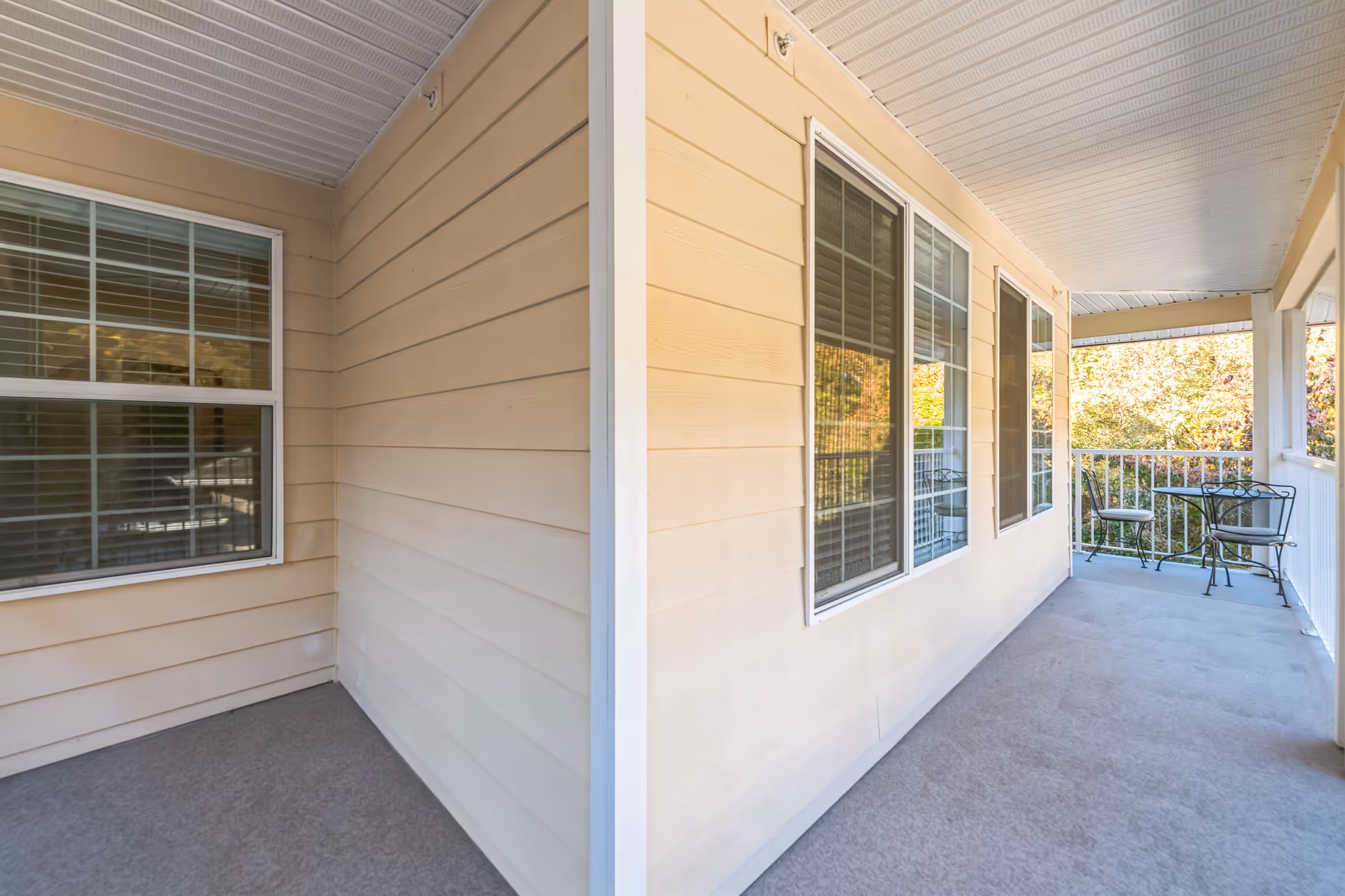 Covered outdoor porch area with beige siding walls, multiple windows with white frames, and a small metal table with two chairs at the far end. The porch has a white ceiling and railing, overlooking greenery outside.