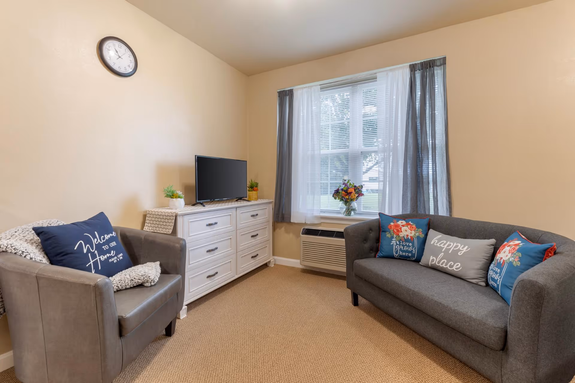 A cozy living room with beige walls and carpet. There is a gray armchair with a 'Welcome to our Home' pillow and a white throw blanket on the left. A white dresser with a small TV and two potted plants sits against the wall. A window with sheer white and gray curtains lets in natural light. In front of the window is a gray sofa with three decorative pillows, two with floral designs and one that says 'happy place'. A clock is mounted on the wall above the armchair.