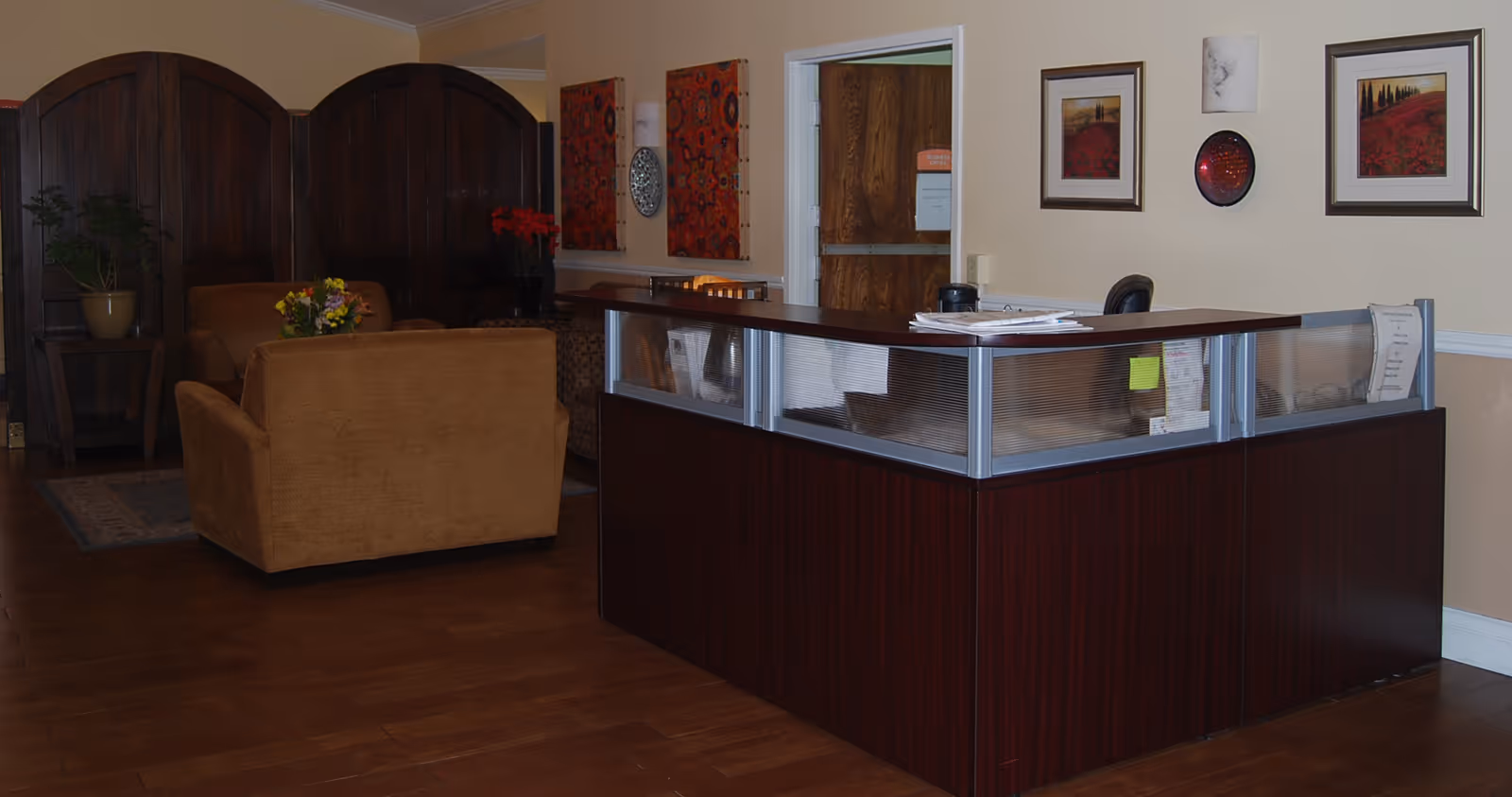 Reception area with a dark wood front desk, a brown couch, a small table with a plant, and decorative artwork on beige walls.