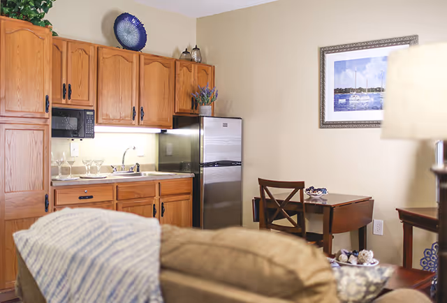 A cozy kitchen area with wooden cabinets, a stainless steel refrigerator, a microwave, and a sink. There are wine glasses on the counter and decorative items on top of the cabinets. A small wooden dining table with two chairs is placed against the wall, which has a framed picture of sailboats on the water. In the foreground, part of a beige couch with a striped blanket is visible.
