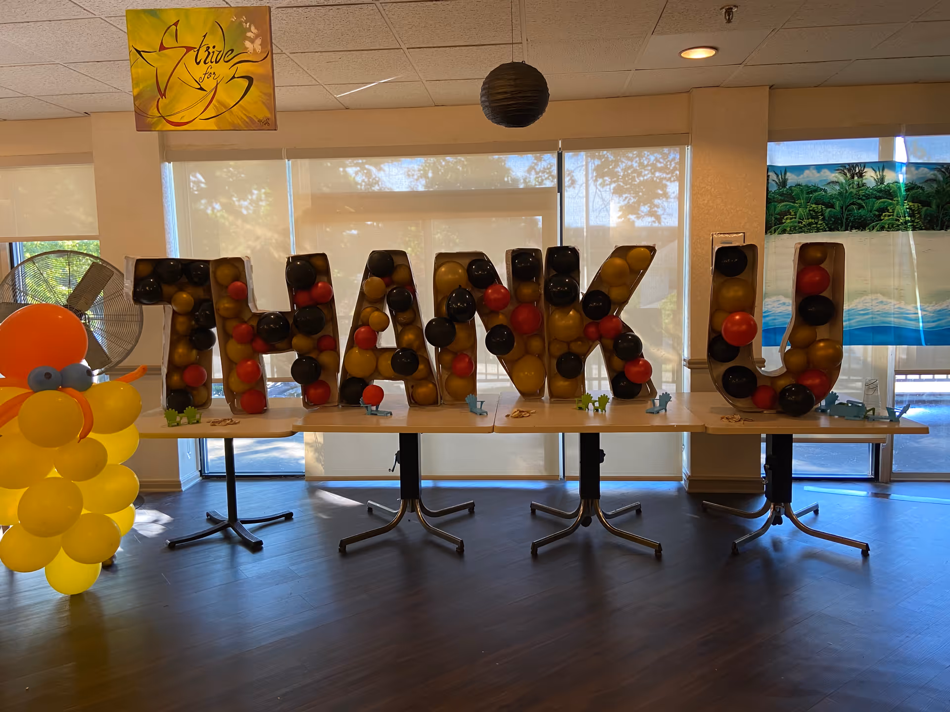 A decorated table inside a room with large windows covered by blinds. On the table are large letters spelling out 'THANK U' filled with red, black, and gold balloons. To the left of the table is a balloon sculpture resembling a yellow duck with an orange beak and blue eyes. A sign hanging from the ceiling reads 'Strive for 5'.