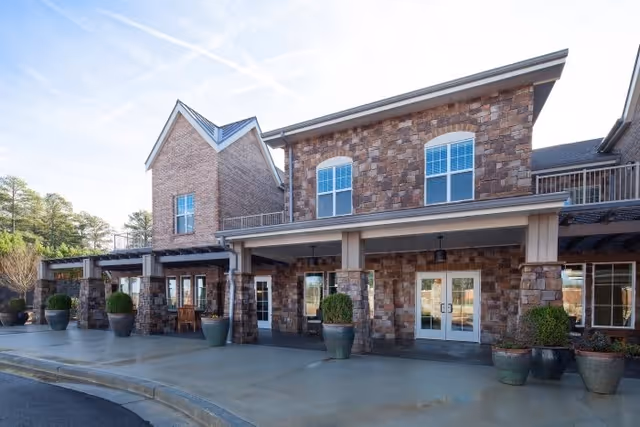 Exterior view of Arbor Terrace Hamilton Mill showing a two-story building with stone and brick facade, large windows, and a covered entrance with potted plants along the walkway.
