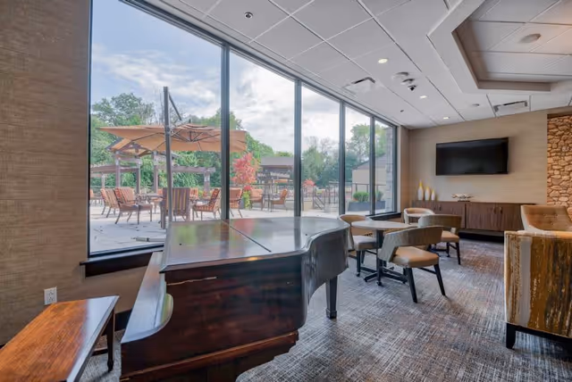Interior view of a lounge area with a grand piano, several chairs, tables, and a large window showing an outdoor patio with umbrellas and seating. The room has a carpeted floor, a wall-mounted TV, and decorative elements including a stone accent wall.