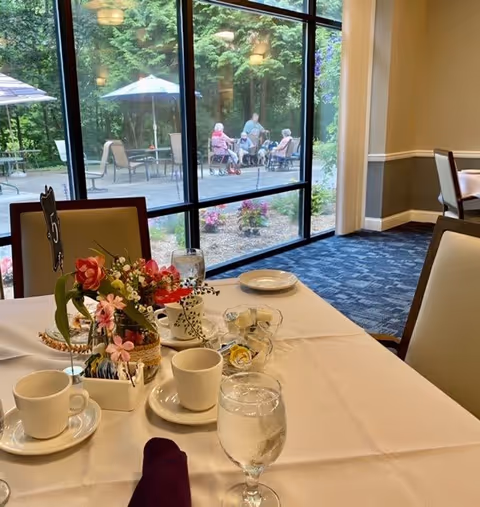Table set for tea or coffee with cups, saucers, a glass of water, a floral centerpiece, and a table number 5 in a dining room. Large windows show an outdoor patio area with elderly people sitting and conversing under umbrellas surrounded by greenery.