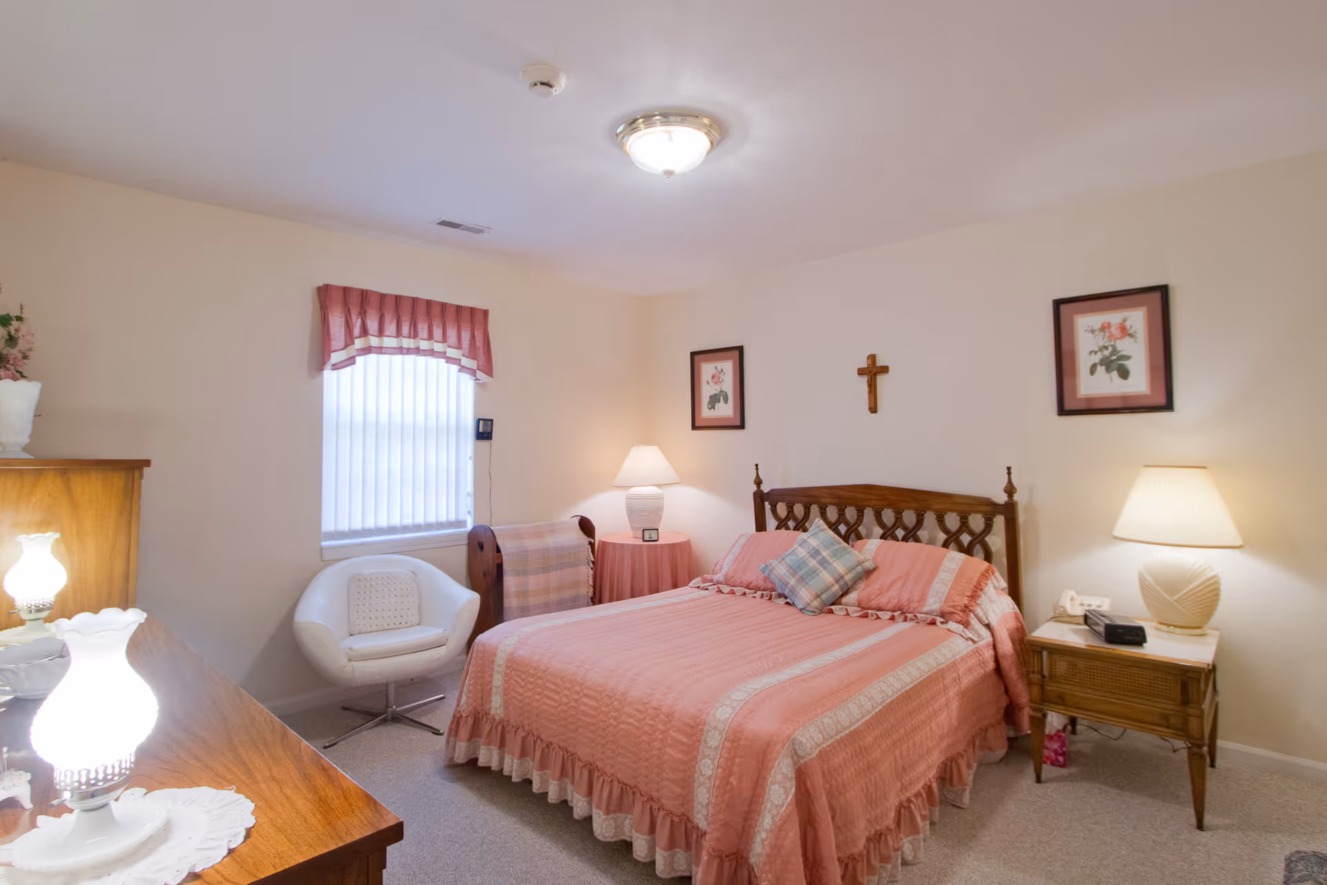 A cozy bedroom with a wooden bed frame and a pink bedspread with matching pillows. The room has two bedside tables with lamps, a white cushioned chair near a window with a pink valance, and framed floral artwork on the walls. A wooden dresser with a lamp is partially visible in the foreground.