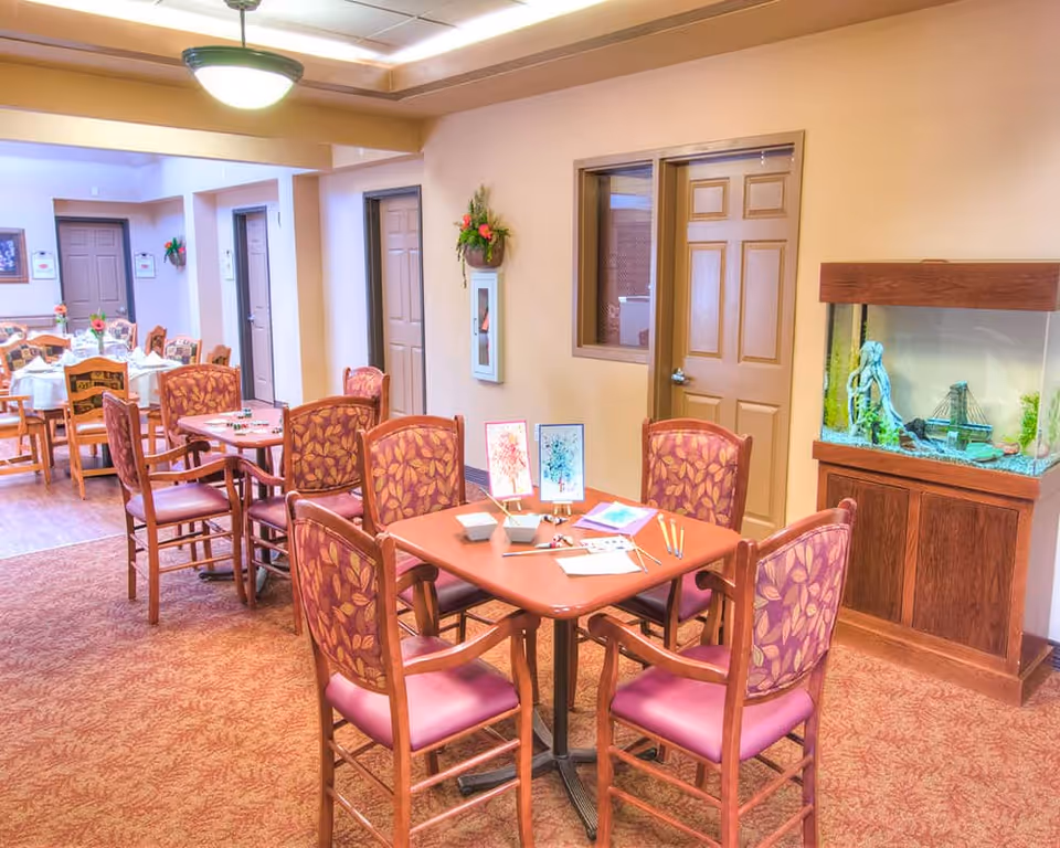 A cozy common area in a senior living facility with multiple tables and chairs arranged for activities and dining. One table in the foreground has art supplies and small paintings displayed. The room features warm lighting, beige walls, a carpeted floor, and a large fish tank on a wooden stand. Several closed doors and wall decorations are visible in the background.