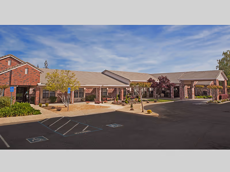 Single-story brick assisted living building with a covered entrance, landscaped courtyard, and empty parking lot with handicap spaces.