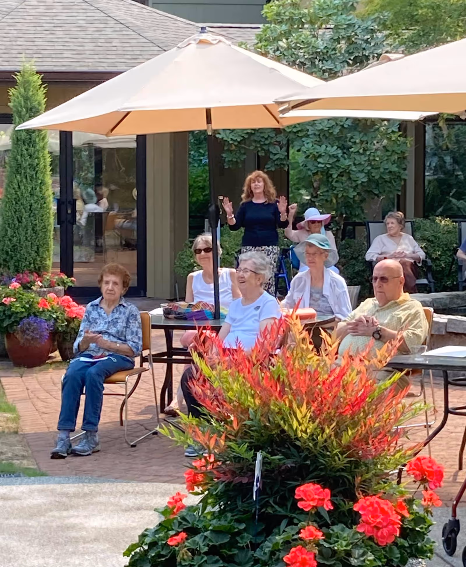 A group of elderly people sitting outdoors under large beige umbrellas in a garden area with colorful flowers and greenery. Some are seated at tables while others sit on chairs, enjoying a sunny day at a senior living facility. A woman stands in the background with her hands raised, possibly speaking or leading an activity.