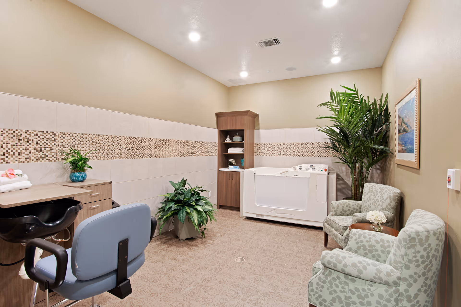 A well-lit room with a walk-in bathtub, two patterned armchairs with a small table between them, a tall potted plant, a cabinet with towels and decorative items, a salon-style chair, and a counter with a small plant and folded towels. The walls have a beige and tiled mosaic design.