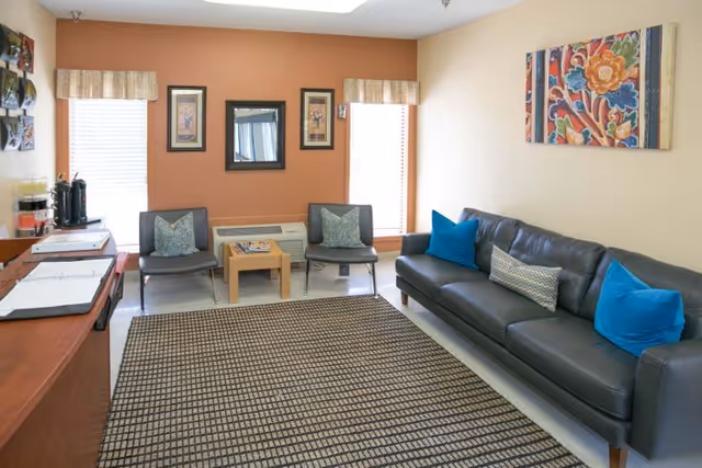 A cozy living room area with a dark leather sofa adorned with blue and patterned throw pillows, two gray chairs with pillows, a small wooden coffee table with magazines, and a patterned area rug. The walls are painted beige and orange, decorated with framed artwork and a colorful floral painting. Two windows with valances allow natural light into the room.