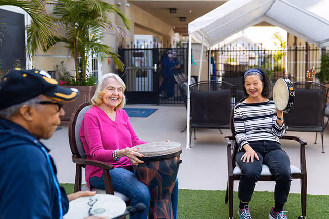 Three elderly individuals sitting outdoors on chairs, playing hand drums and a tambourine, smiling and enjoying a musical activity in a courtyard area with a white canopy and gated entrance in the background.