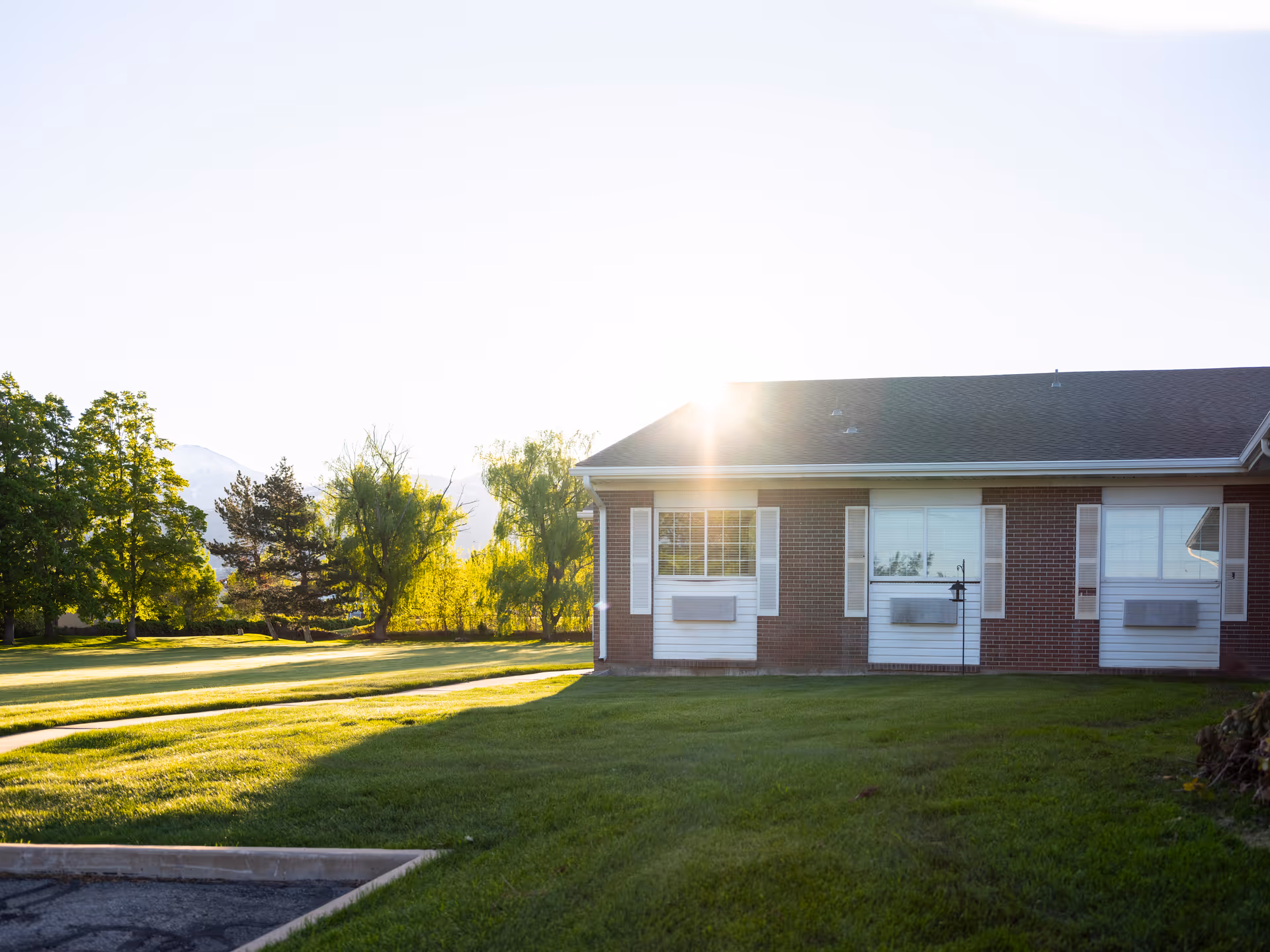 Exterior view of a single-story brick building with white window shutters and air conditioning units below the windows, surrounded by a well-maintained green lawn and trees with the sun shining brightly in the background.