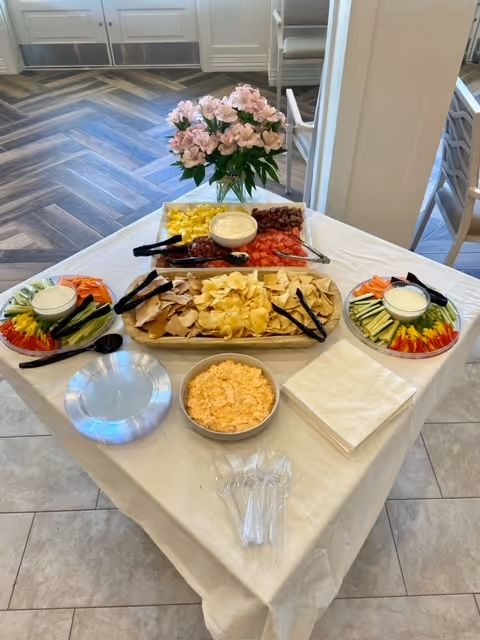 A table set with a variety of snacks including trays of fresh vegetables with dip, a platter of chips with pita bread, a bowl of cheese spread, and a tray of cut fruits such as pineapple, grapes, and watermelon. The table is covered with a white tablecloth and has plastic utensils, napkins, and plates. A vase with pink flowers is placed at the back of the table. The setting appears to be indoors with herringbone patterned wooden flooring and chairs in the background.