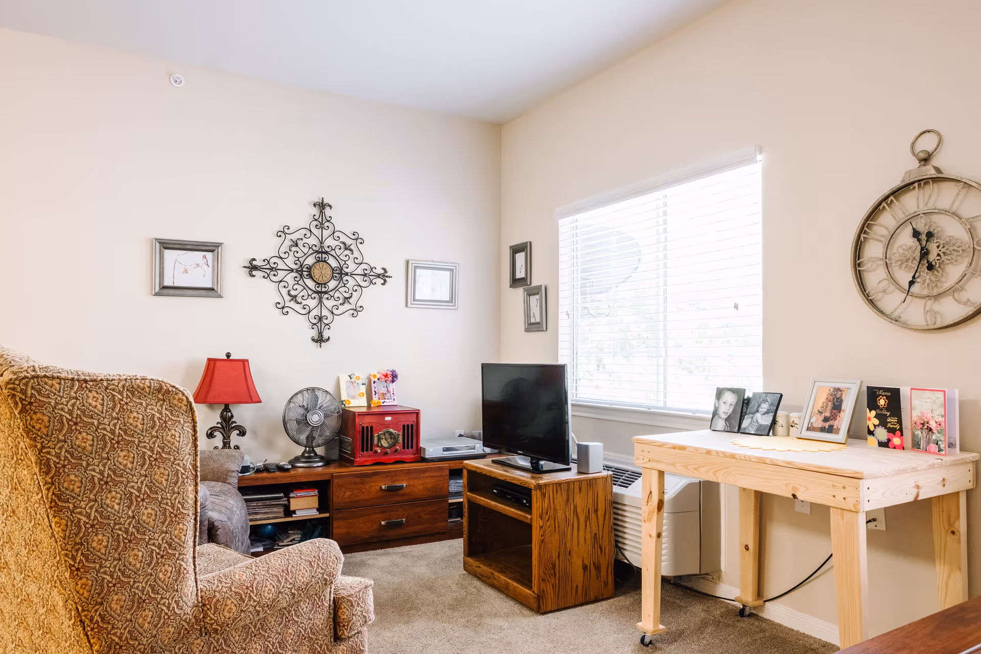 A cozy living room with a patterned armchair, a wooden TV stand holding a flat-screen television, and a wooden side table with framed photos and decorative items. The walls are decorated with a large ornate wall clock, a decorative metal wall piece, and framed pictures. A window with white blinds lets in natural light.