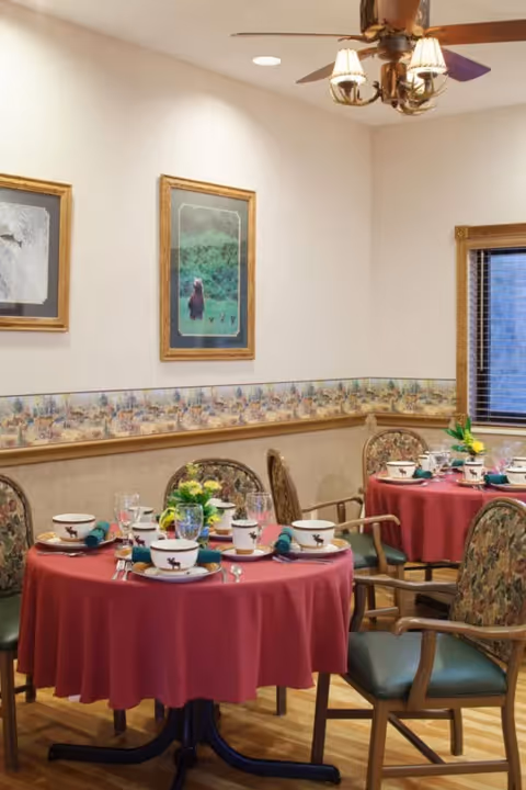 A dining room with round tables covered in red tablecloths, set with cups, plates, utensils, and napkins. Each table has a small floral centerpiece. The room has floral-patterned chairs, framed artwork on the walls, a ceiling fan with lights, and a window with blinds.