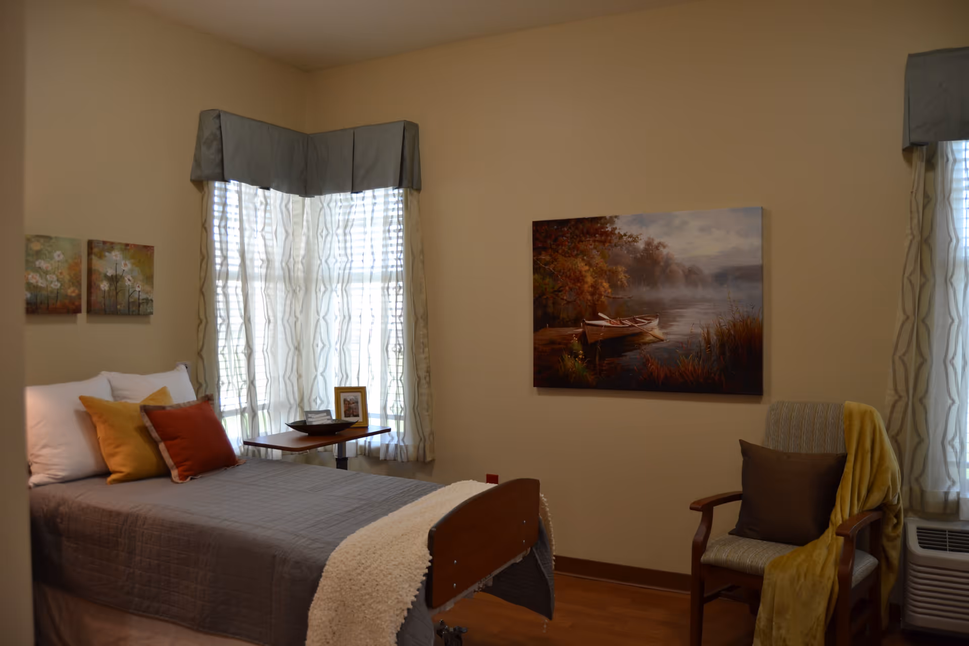 A cozy bedroom in an assisted living facility featuring a single bed with gray bedding and multiple pillows in white, yellow, and red. There is a small table next to the bed with a framed photo and a decorative bowl. The room has two windows with sheer curtains and gray valances. On the wall above a wooden chair draped with a yellow blanket and a brown pillow, there is a large painting of a boat on a calm lake surrounded by autumn trees.