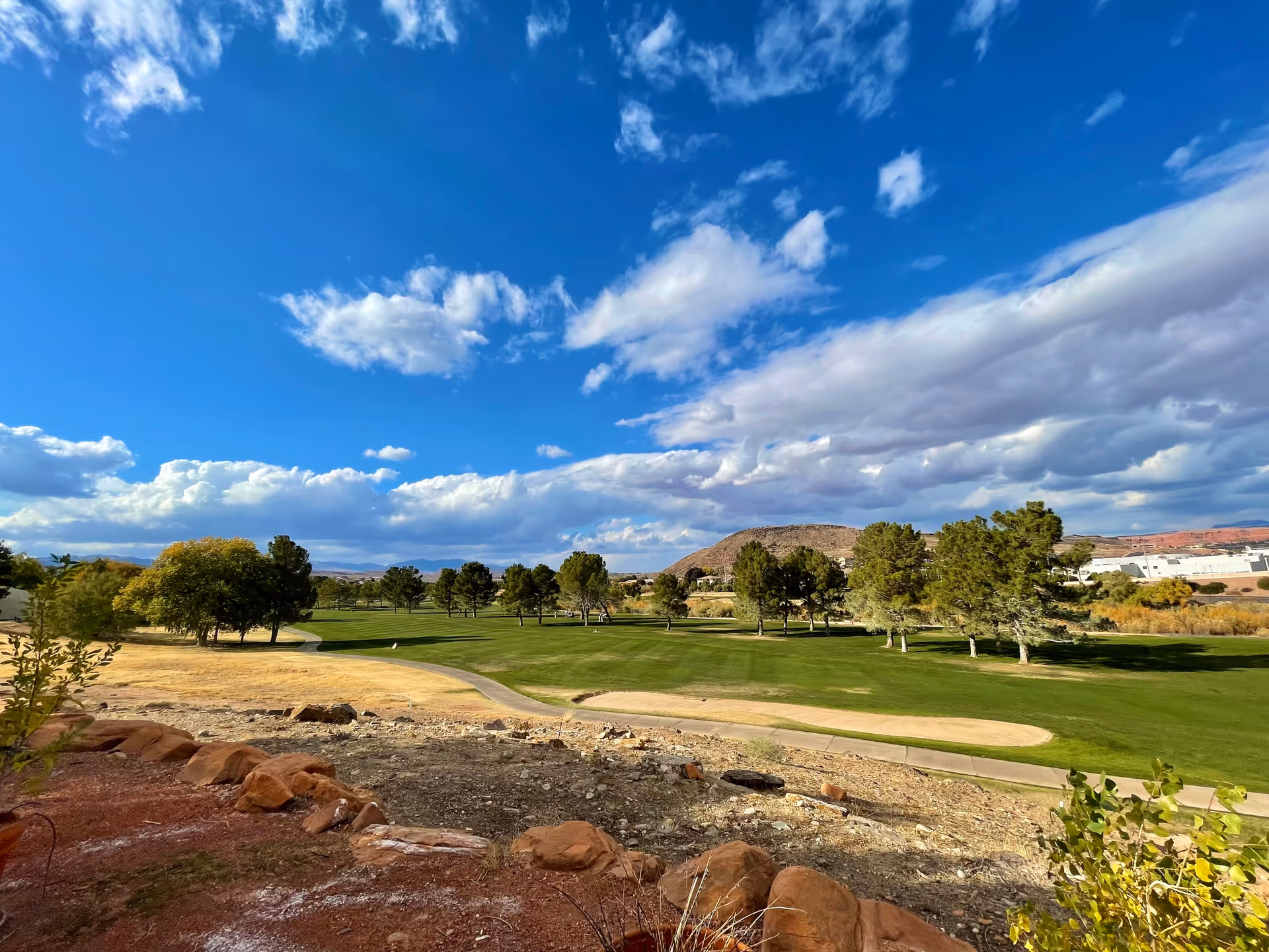A scenic outdoor view of a golf course with green grass, trees, a sand trap, and a paved path under a blue sky with scattered clouds. In the background, there are hills and some buildings.
