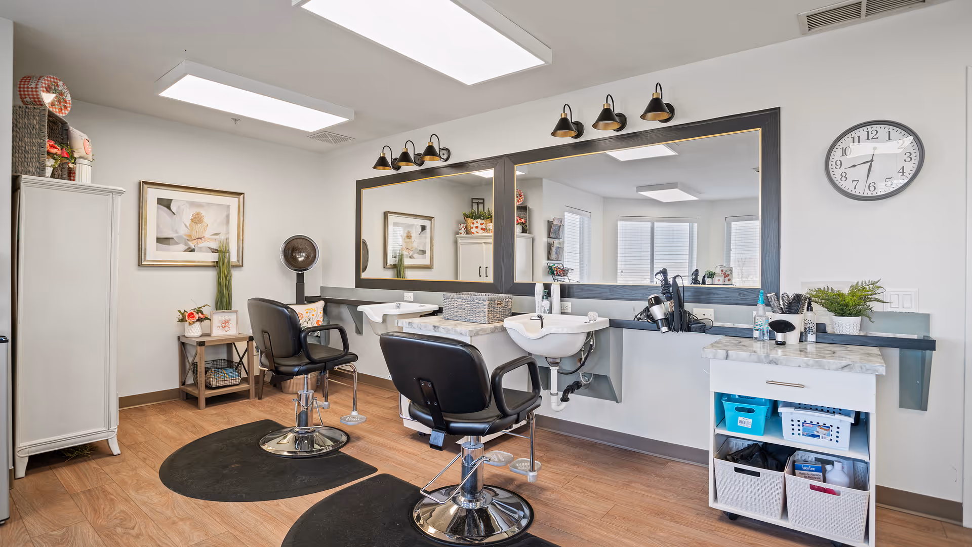 Interior view of a hair salon area in a senior living facility with two black salon chairs in front of large mirrors, two hair washing sinks, a white cabinet with hair care products, a clock on the wall, and decorative plants and artwork.