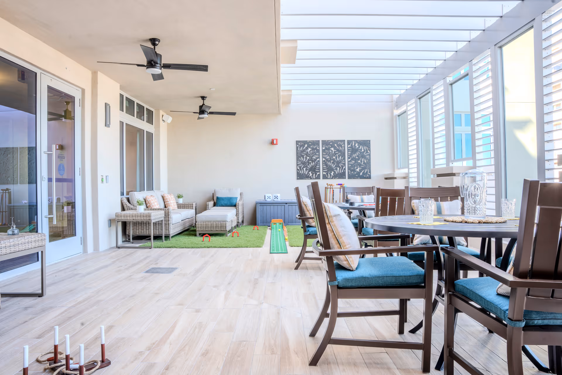 A bright and airy indoor lounge area with large windows and ceiling fans. The space features comfortable wicker seating with cushions, a small putting green, and a round dining table with chairs that have blue cushions. The floor is light wood, and there are decorative wall panels on the far wall.
