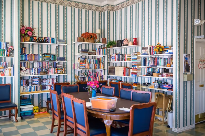 A bright communal room with bookshelves lining striped wallpaper and a large table surrounded by blue-upholstered chairs.