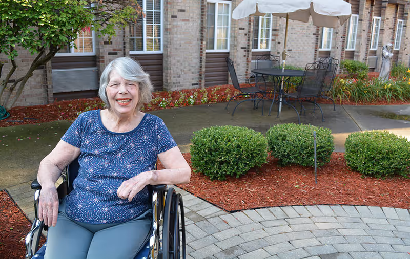 An elderly woman with gray hair sitting in a wheelchair outdoors in a garden area with bushes, a paved walkway, and a patio table with chairs and an umbrella in the background. She is smiling and wearing a blue patterned shirt and gray pants.