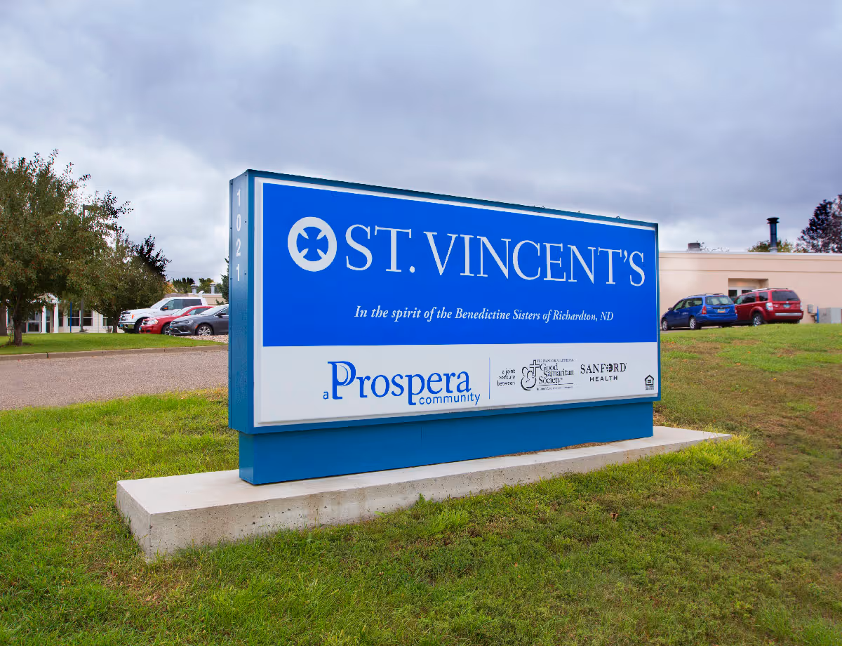 Outdoor view of a large blue and white sign for St. Vincent's, a Prospera community, located on a grassy area with a parking lot and building in the background under a cloudy sky.