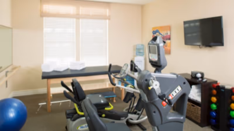 A small fitness room with exercise equipment including a recumbent bike and an elliptical machine. There is a blue exercise ball on the floor, a massage or treatment table with folded towels, a water dispenser, a wall-mounted TV, and a rack holding colorful dumbbells. The room has beige walls and two windows with blinds.