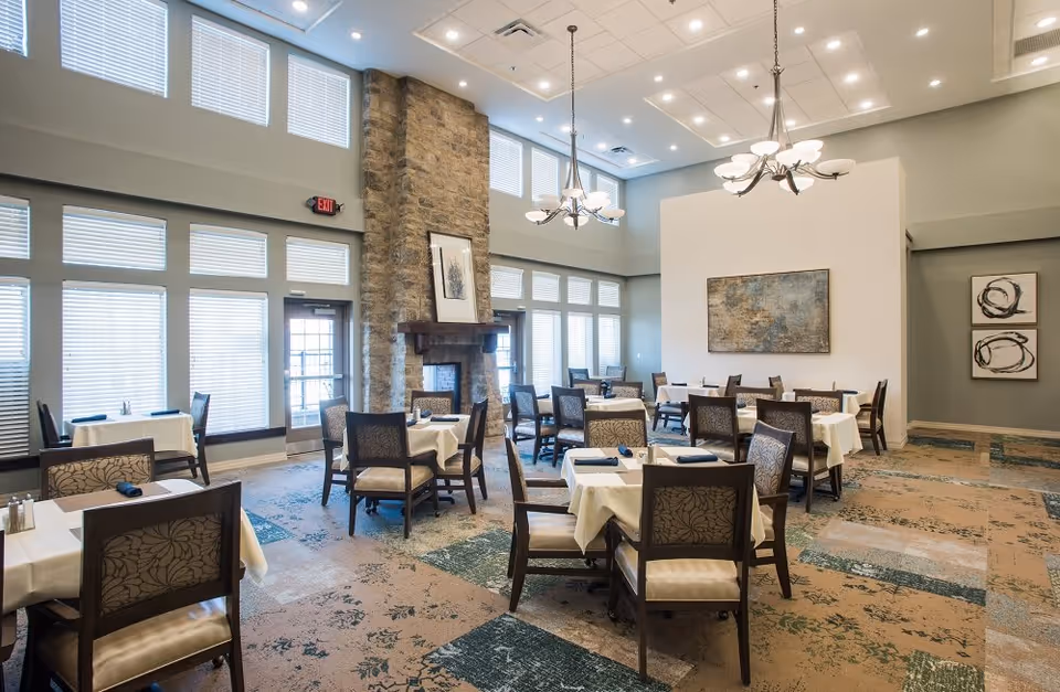 A spacious dining room with multiple tables covered in white tablecloths and set with dark napkins. The room features large windows with blinds, a stone fireplace, modern chandeliers, and abstract artwork on the walls. The carpet has a patterned design in shades of green and beige.