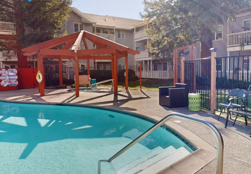 Outdoor swimming pool area at Cogir of Rohnert Park with a curved pool, metal handrail, a wooden pergola, seating including chairs and a bench, surrounded by a black metal fence and multi-story residential buildings in the background.